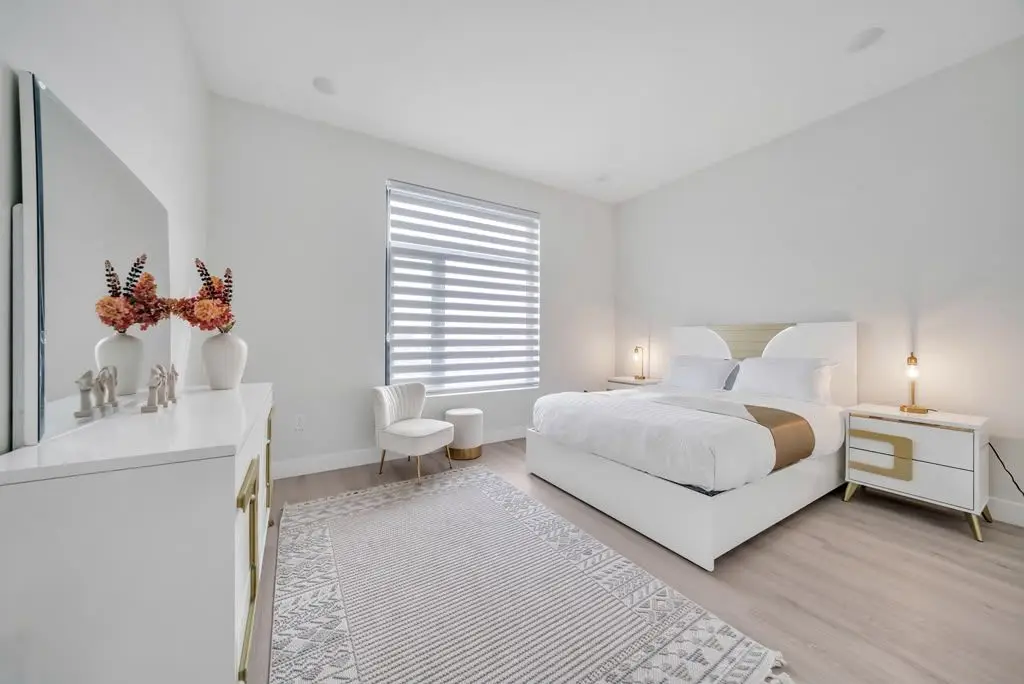 Bedroom with white upholstered bed, dresser, accent chair and a window with zebra blinds, Residence 106