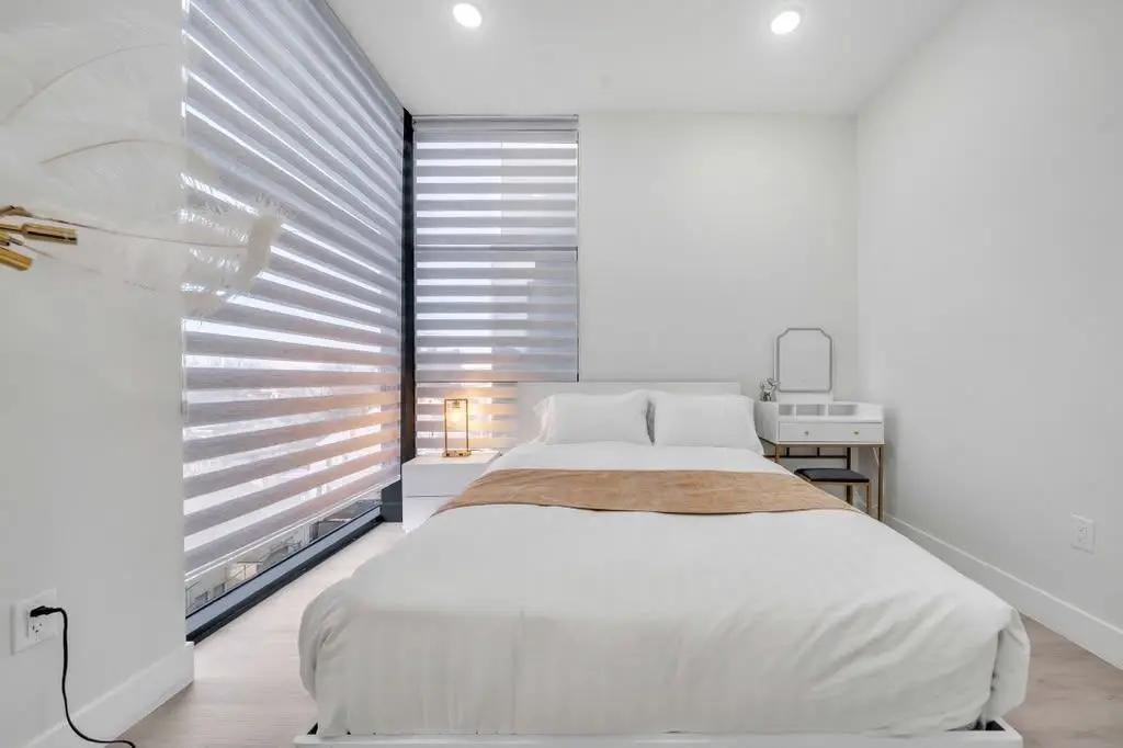 Bedroom with white bed, tan throw and a large corner window with blinds, Residence 407